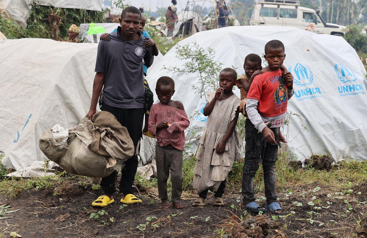 A man stands in front of UNHCR tents, with a young child on his back and four other children standing beside him