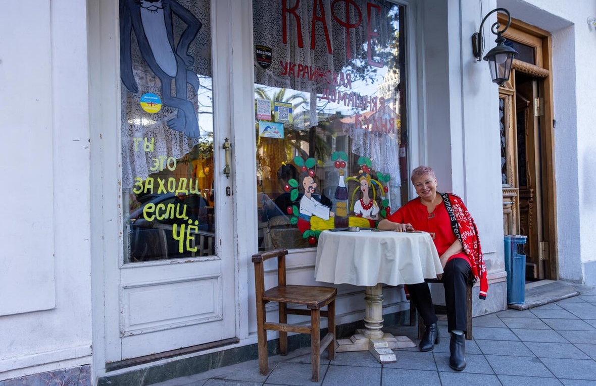 A smiling woman seated at a table in front of a café with colorful paintings on the windows and doors.