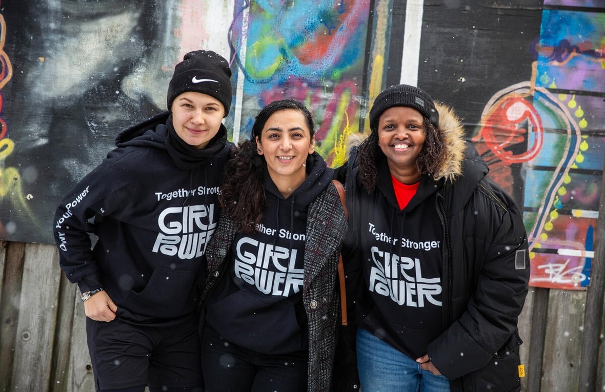 Three women wearing 'Girl Power' jumpers smile at the camera, their arms around each other, in front of a colorful building
