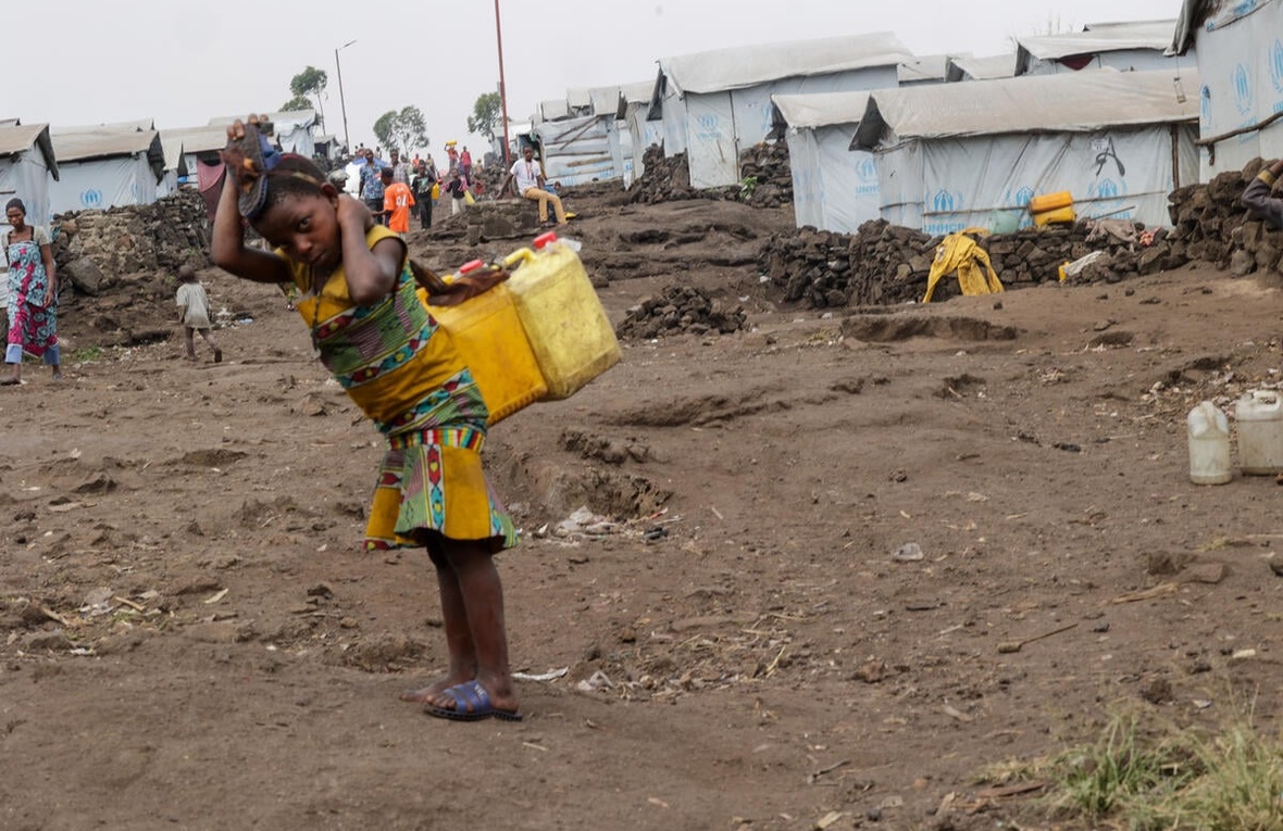 A small girl carries two yellow cannisters of water in a settlement for displaced people.