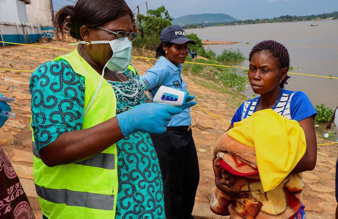 A woman wearing a mask prepares to take another woman's temperature next to a river, while a police officer stands nearby.