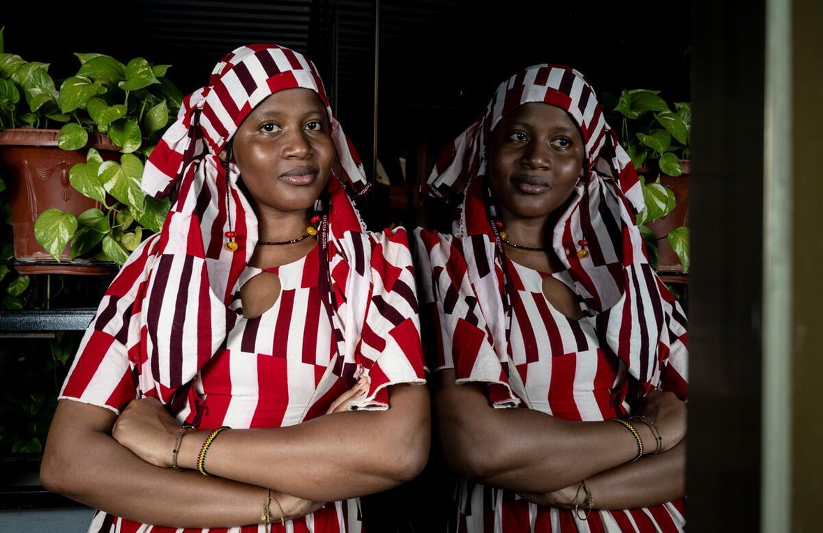 A woman in a red and white striped dress and headscarf leans against a window in which she is reflected