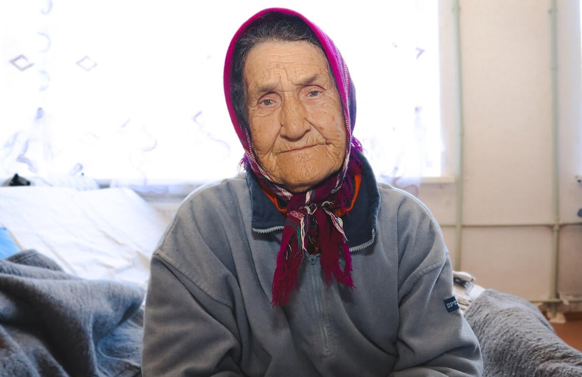 Portrait of older woman sitting on a bed, wearing scarf tied under her chin.