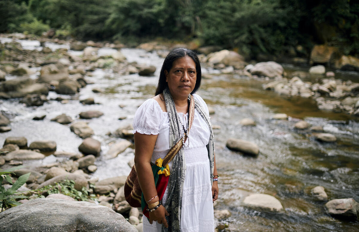 A woman wearing a long white dress with a colourful woven bag on her shoulder stands on the bank of a shallow rocky river  