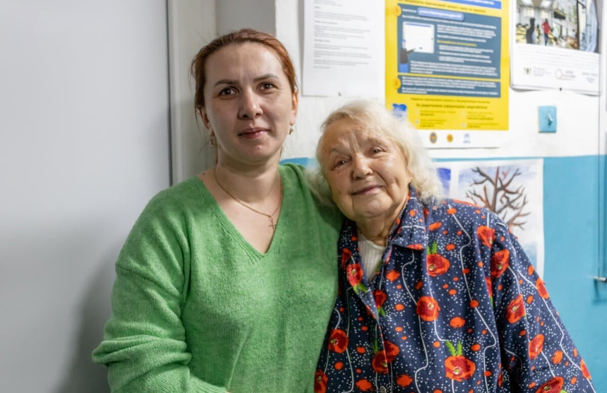 A younger woman and an older woman stand together, arms around each other.