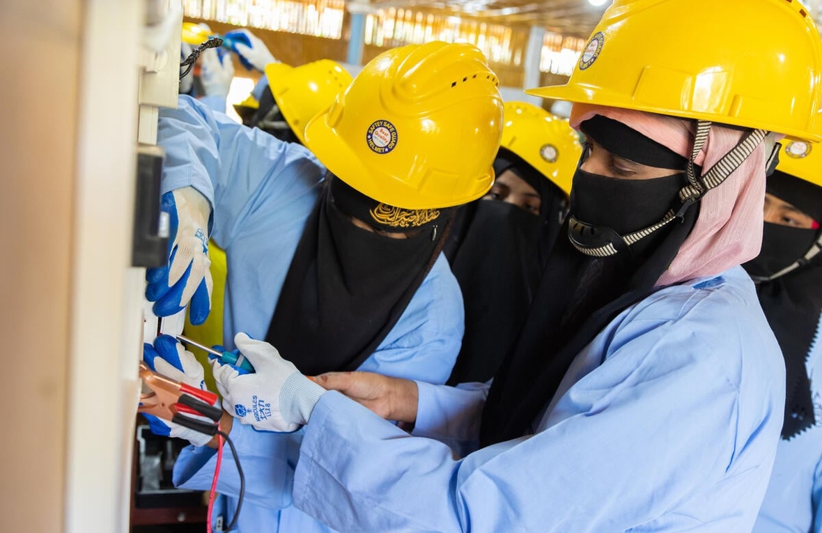 Two women wearing niqabs and yellow hardhats work together to screw a switch to a wall while other women look on.