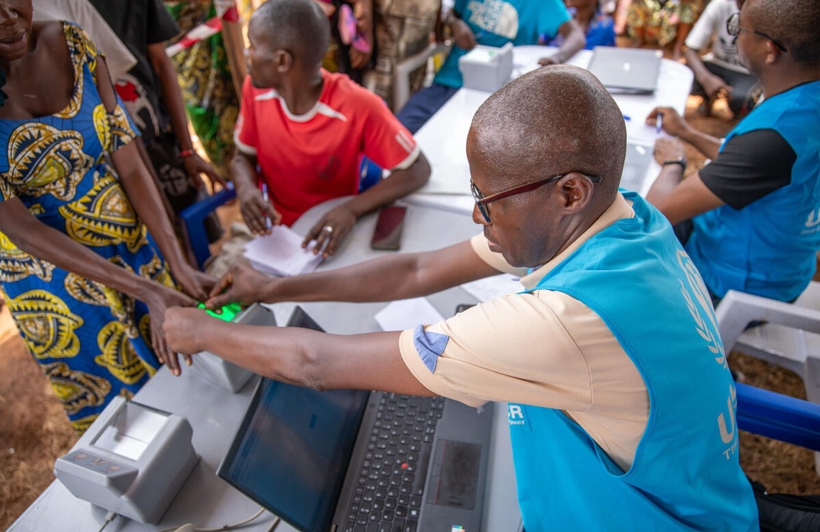 A UNHCR staff member seated at a desk with a laptop assists a woman to scan her thumbprints 