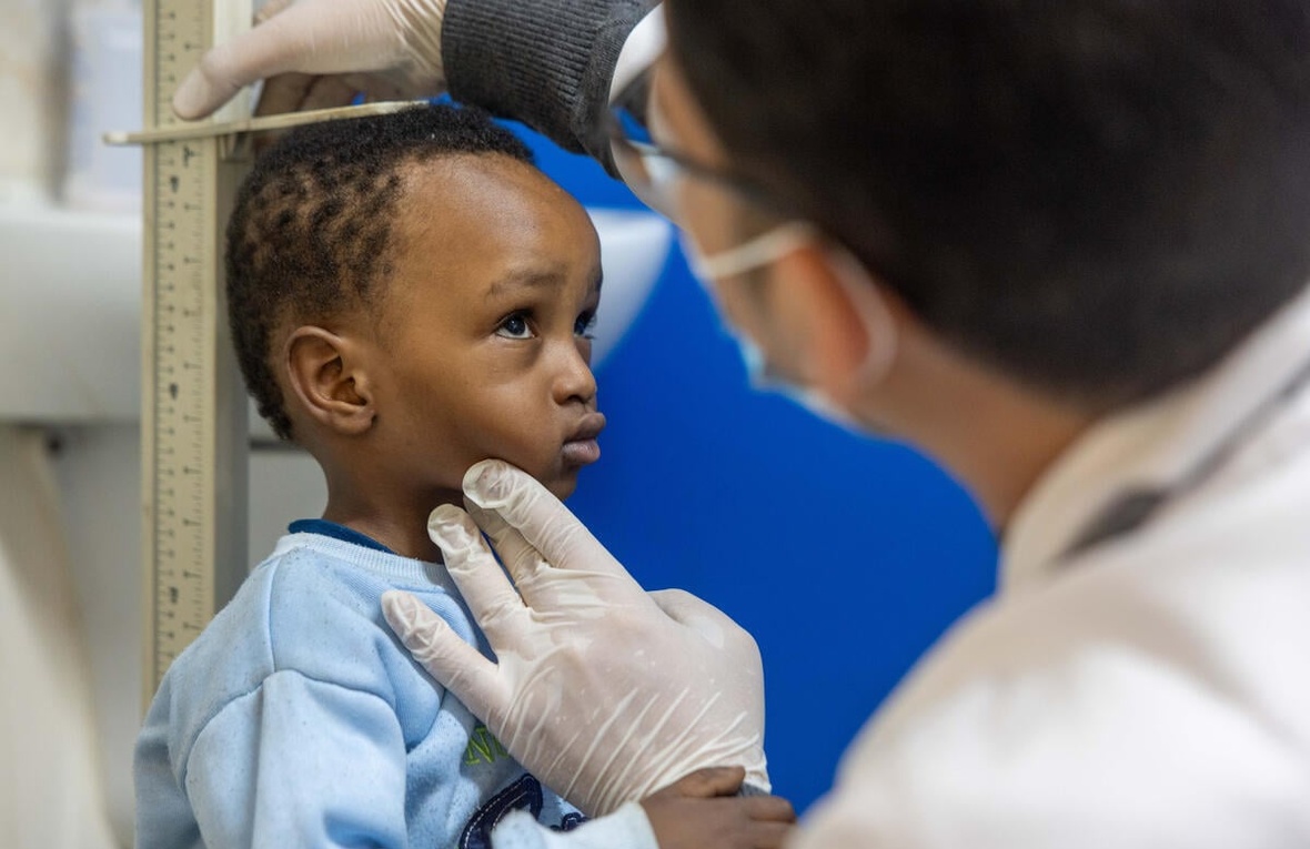 A young boy in a blue sweater stands against a height rule as a man wearing a surgical mask and gloves measures his height.
