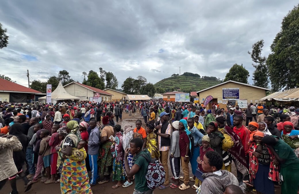 Large groups of Congolese refugees gather outside some buildings and shelters with trees and hills behind them.