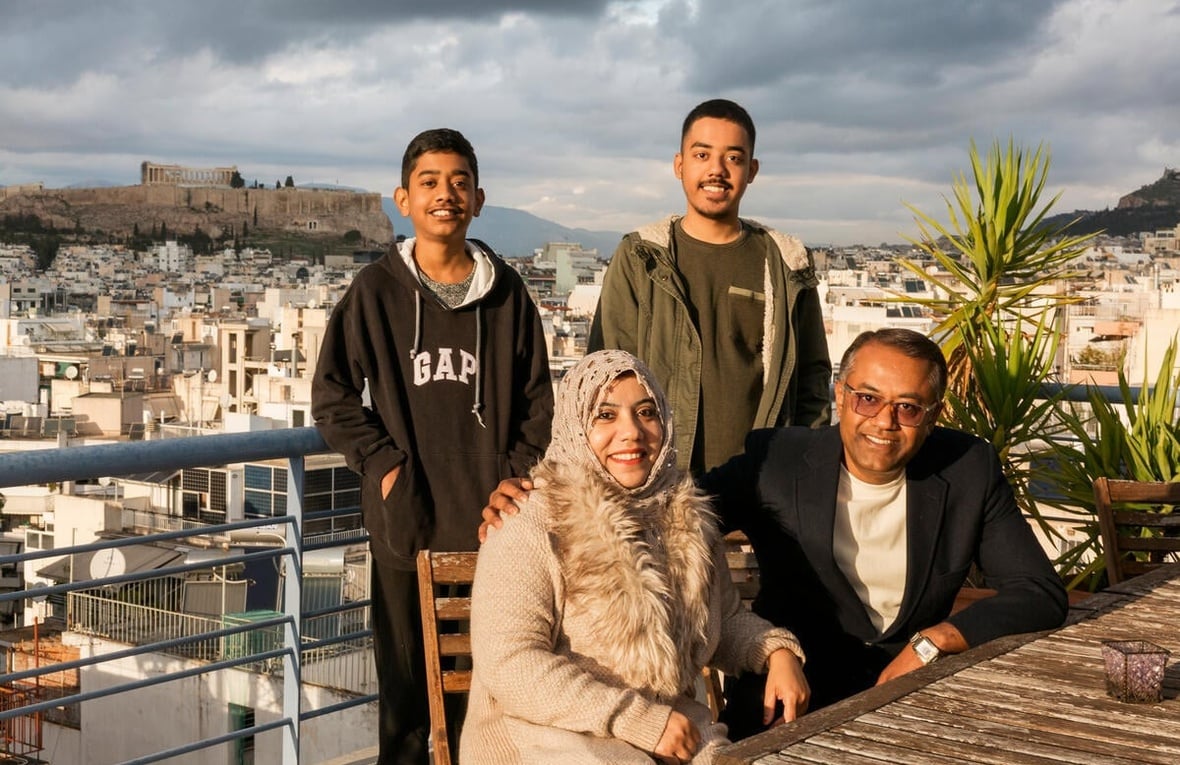 A middle-aged couple sit on a balcony with their two sons standing behind them, with a view of Athens behind them. 