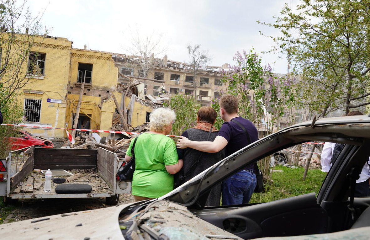 Three people pictured from behind, a damaged car behind them, as they observe a damaged building