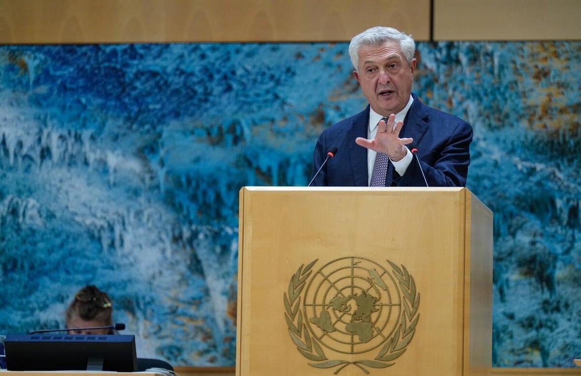 A man wearing a suit makes a hand gesture while speaking at a wooden lectern bearing the UN logo