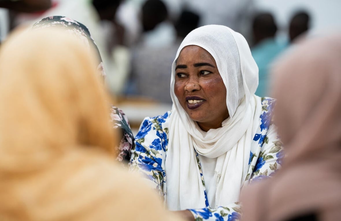 A smiling woman in a white headscarf and blue floral dress sits at a table with other women