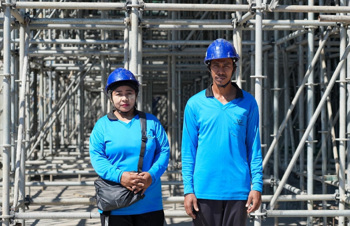 A woman and a man wearing matching blue shirts and hard hats stand outside in front of scaffolding