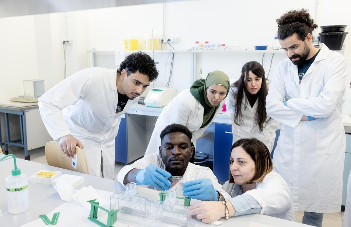 A group of people wearing lab coats gather round as a student and professor carry out an experiement in a laboratory 