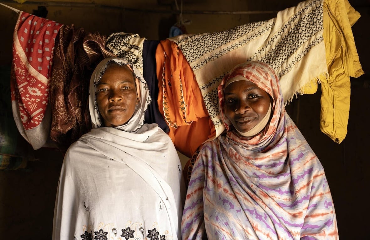 Two women stand next to each other inside a room with washing hanging on a line behind them