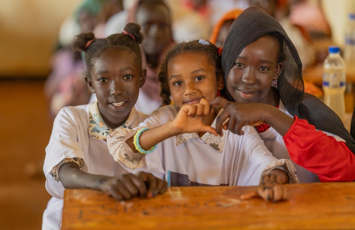 Three Sudanese refugee pupils sit at a desk, smiling, two of them holding their hands together to make a heart shape.