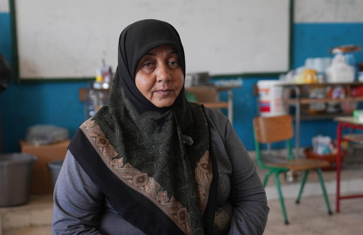 A woman in a headscarf sits in a school classroom with household items on desks in the background