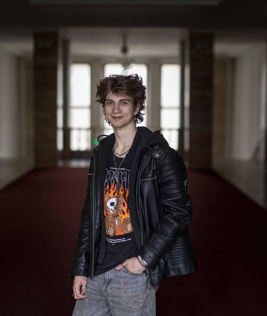 A young man stands smiling indoors, hand in one pocket, with windows behind him