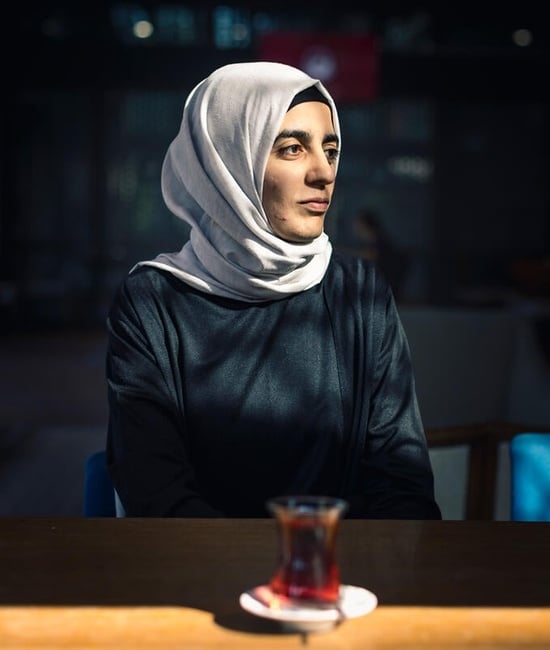 Aicha is surrounded by shadows with her face lit up by the sun as she looks off to the side, a cup of tea on a table in front of her