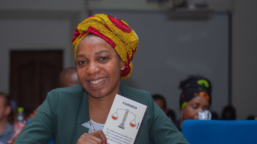 A smiling woman sits at a desk holding up a card that says 'Fairness, justice: You believe that all people have value. You approach situations with an unbiased mindset and treat everyone with respect.'