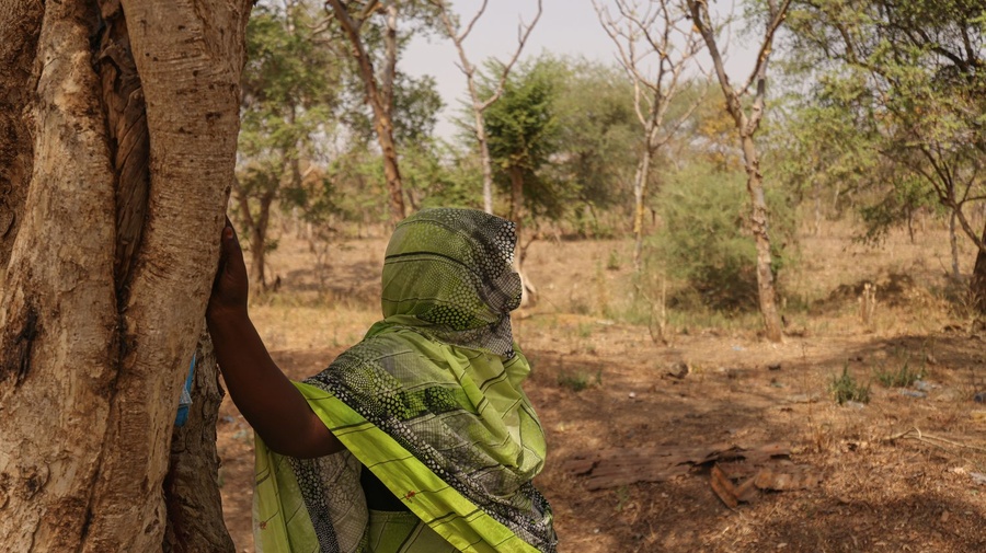 A woman leans against a tree looking out into the scrubland.