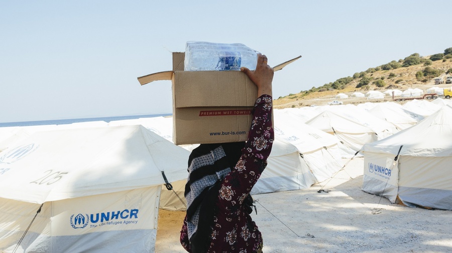 A women carrying water bottles on her head in front of UNHCR tarpaulins