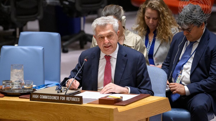 A man in a suit sits at a desk speaking into a microphone with other people seated behind him.