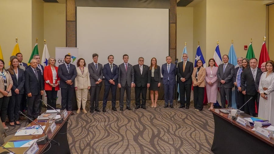 Un grupo de 18 hombres y 11 mujeres con vestimenta formal, de pie en una sala de juntas, detrás de ellos se encuentran 13 banderas coloridas de diferentes países
