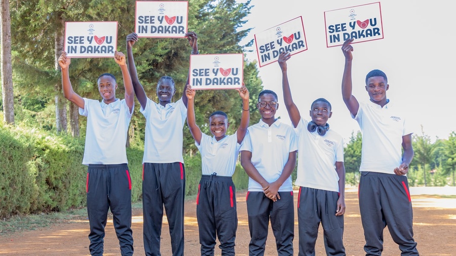 Six smiling young athletes wearing training shoes and clothes hold up signs reading 'see you in Dakar' on a wide dusty road 
