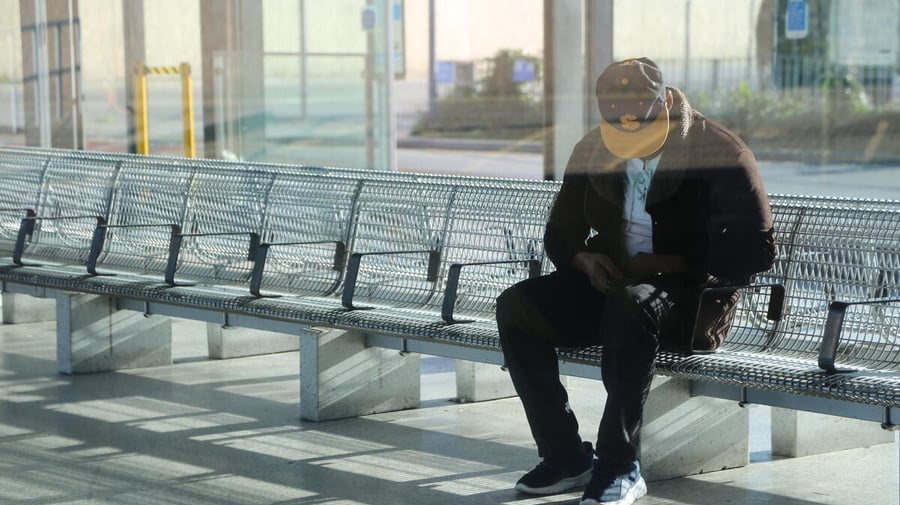 A man sits on metal chairs in a public space in Southeast London, his face obscured by the brim of a cap.