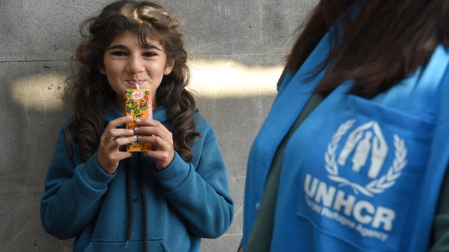 A young refugee girl drinks from a juice box while standing beside a UNHCR staff member.