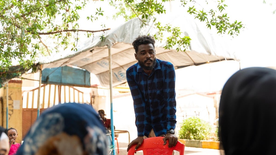 A young man stands in front of a group while leaning on a chair in the courtyard of a school.