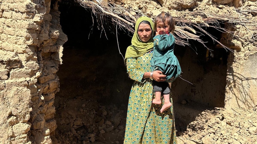 A woman carrying a young girl stands in front of a partially collapsed mud-brick building.