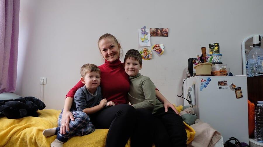 A woman and her two children are pictured sitting together on a bed, arms around each other.