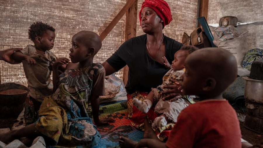 A woman sits cradling an infant inside a shelter with three other young children sitting around her.