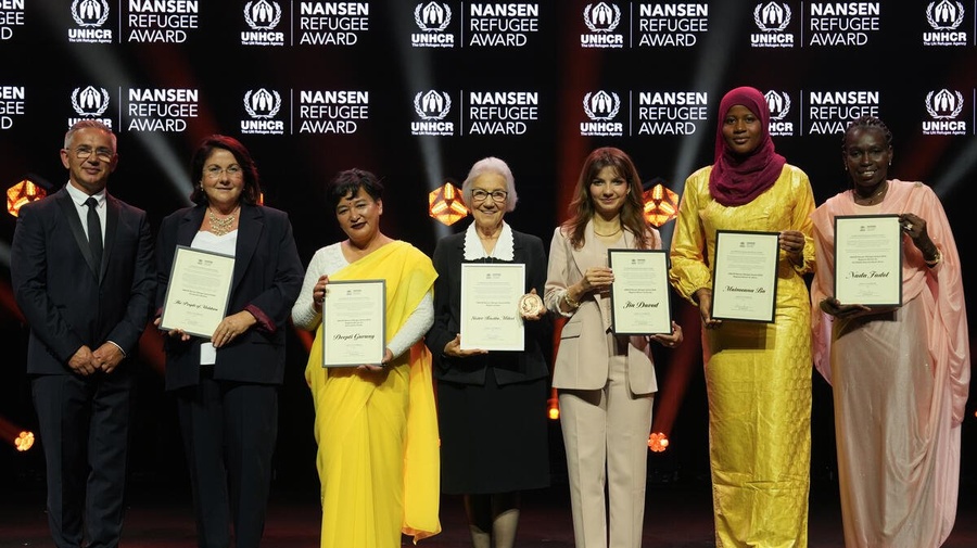 Seven people pose for a photo on a stage. They're holding framed certificates.