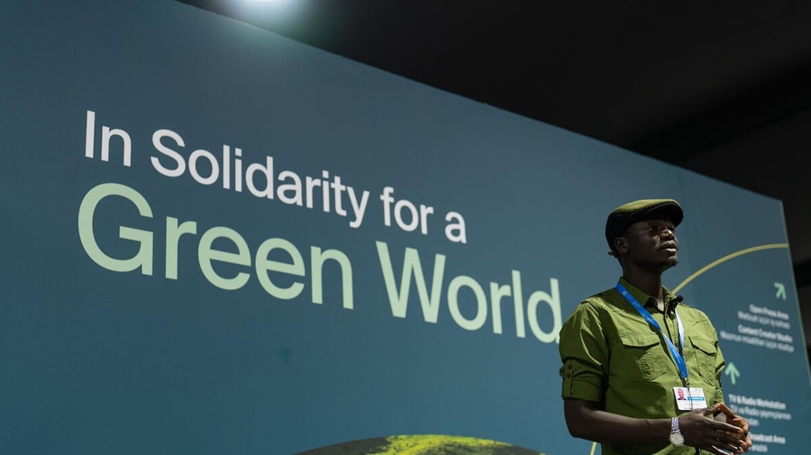 Opira looks into the distance, wearing a lanyard, in front of a huge banner which says 'In Solidarity for a Green World'