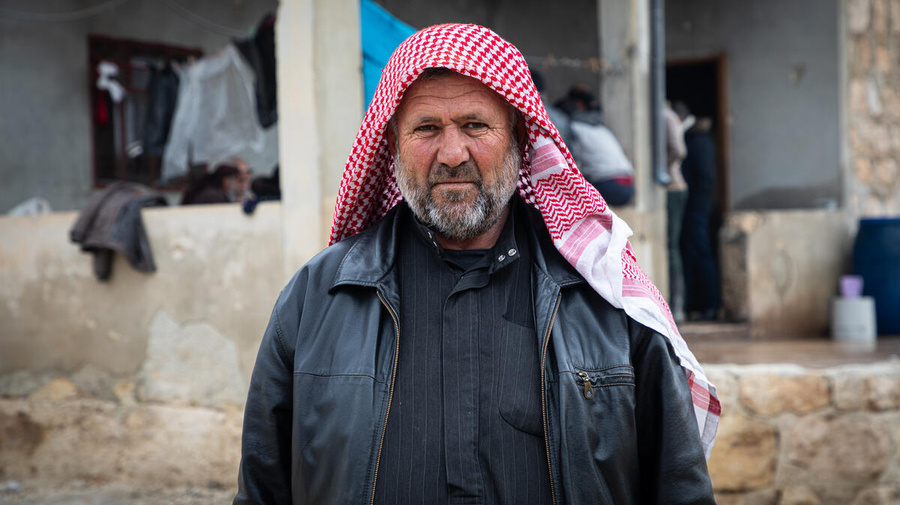 A man wearing a red headscarf and leather jacket stands in front of a building.
