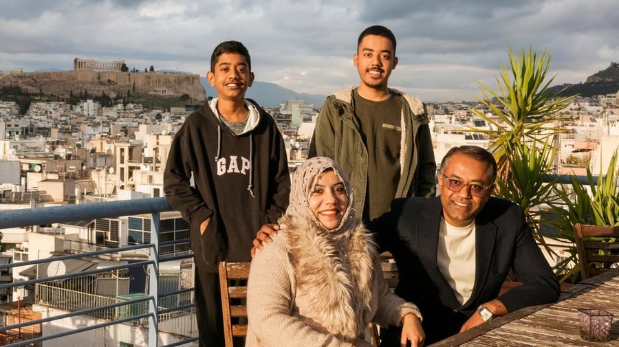 A middle-aged couple sit on a balcony with their two sons standing behind them, with a view of Athens behind them. 