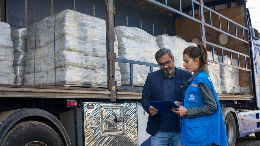 A woman in jeans and a UNHCR vest and a man in jeans and a blazer stand together looking at a clipboard in front of a truck full of supplies