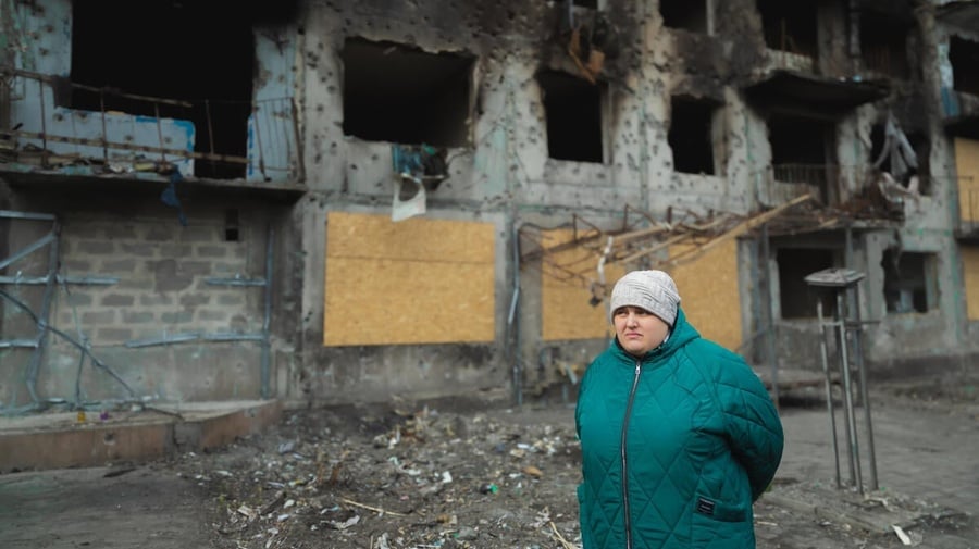 A woman in a winter jacket stands in front of a damaged building.