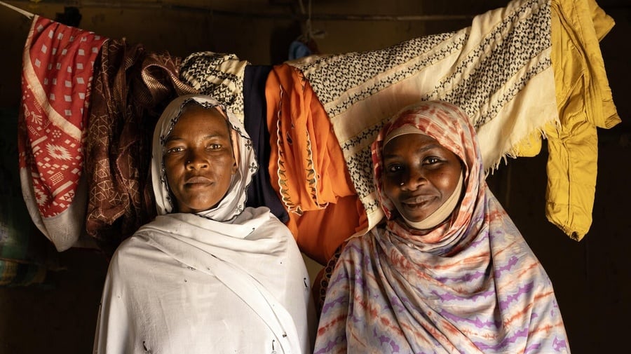Two women stand next to each other inside a room with washing hanging on a line behind them