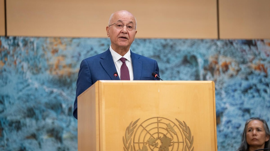A man in a suit and tie speaks from behind a wooden lectern emblazoned with the UN logo