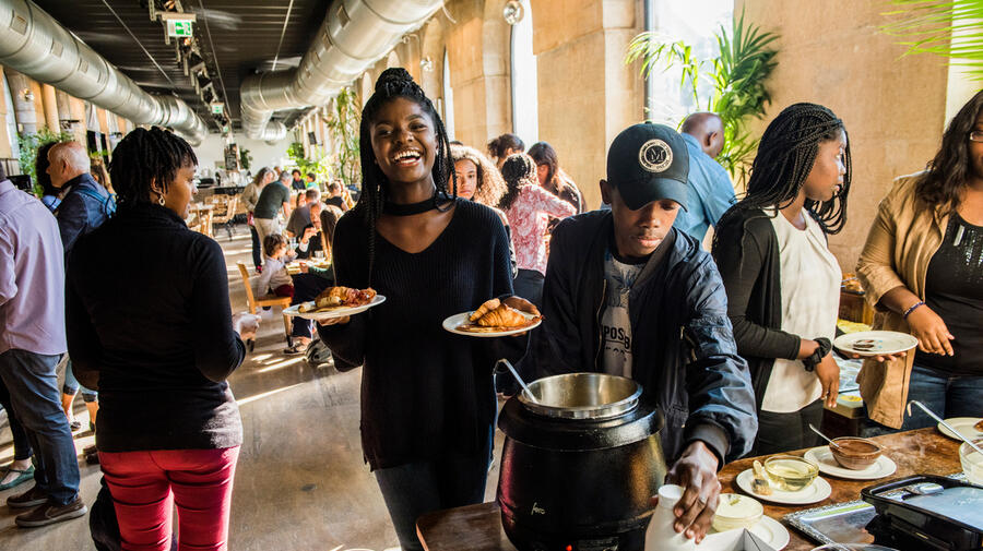 A women, carrying plates of food, smiles at the camera. Other people around her select food from a buffet table in a restaurant.