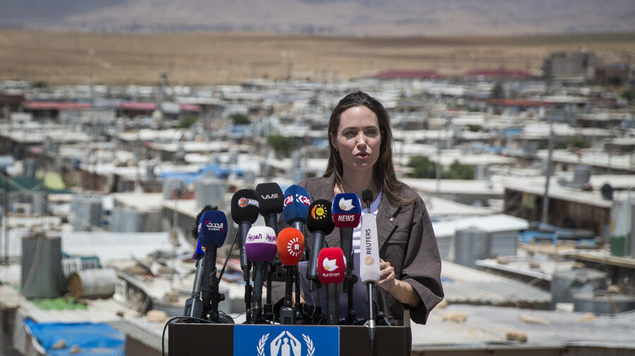 Angelina Jolie stands at a podium covered with news microphones, with Domiz refugee camp in the background