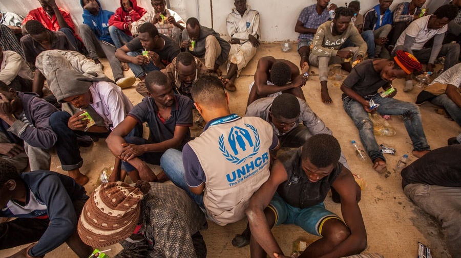 A group of disembarked refugees and migrants sit on the floor, joined by a UNHCR staff member wearing a tan UNHCR vest with blue logo.